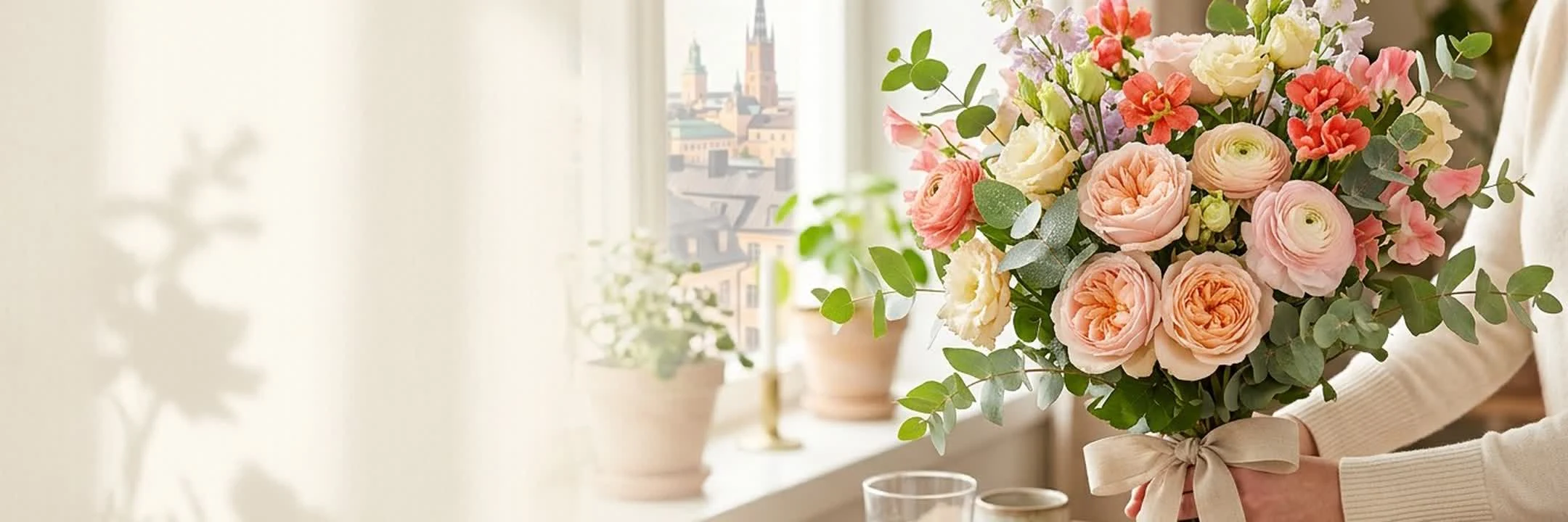 En bukett med persikofärgade rosor och ranunkler vid ett fönster med utsikt över Stockholm.